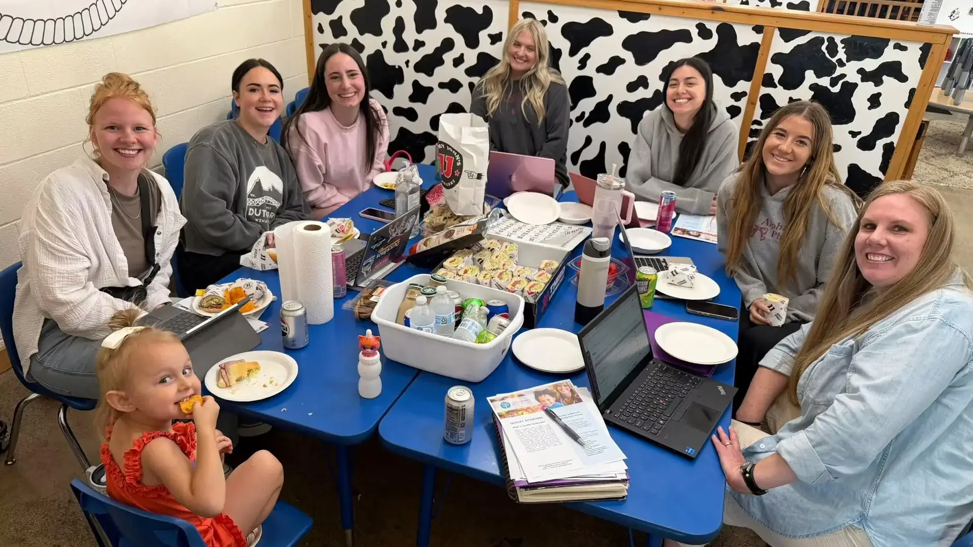 The adult staff at Little Buckaroos Childcare and Preschool sit with a child and IdahoSTARS quality consultant Layna Franklin at a table with food .