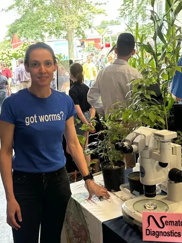 A woman stands by table at an outdoor gathering with laboratory equipment and plants on it.