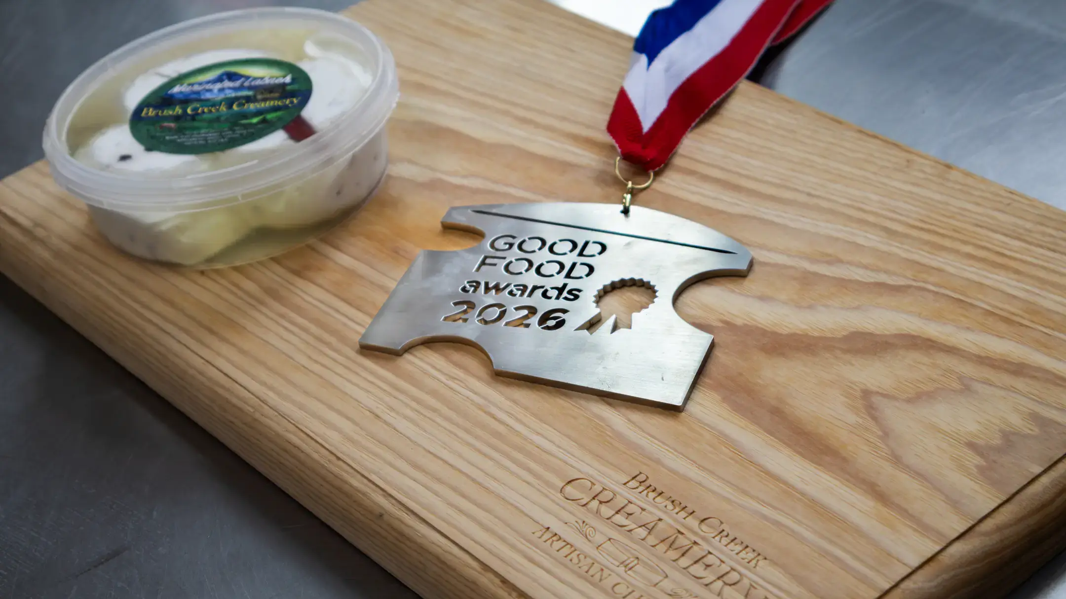 A container of cheese next to a 2026 Good Food Awards medal on a wooden board labeled "Brush Creek Creamery Artisan Cheese."
