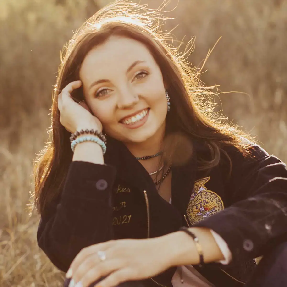 A person smiling outdoors with sunlight highlighting their hair, sitting in a natural setting.