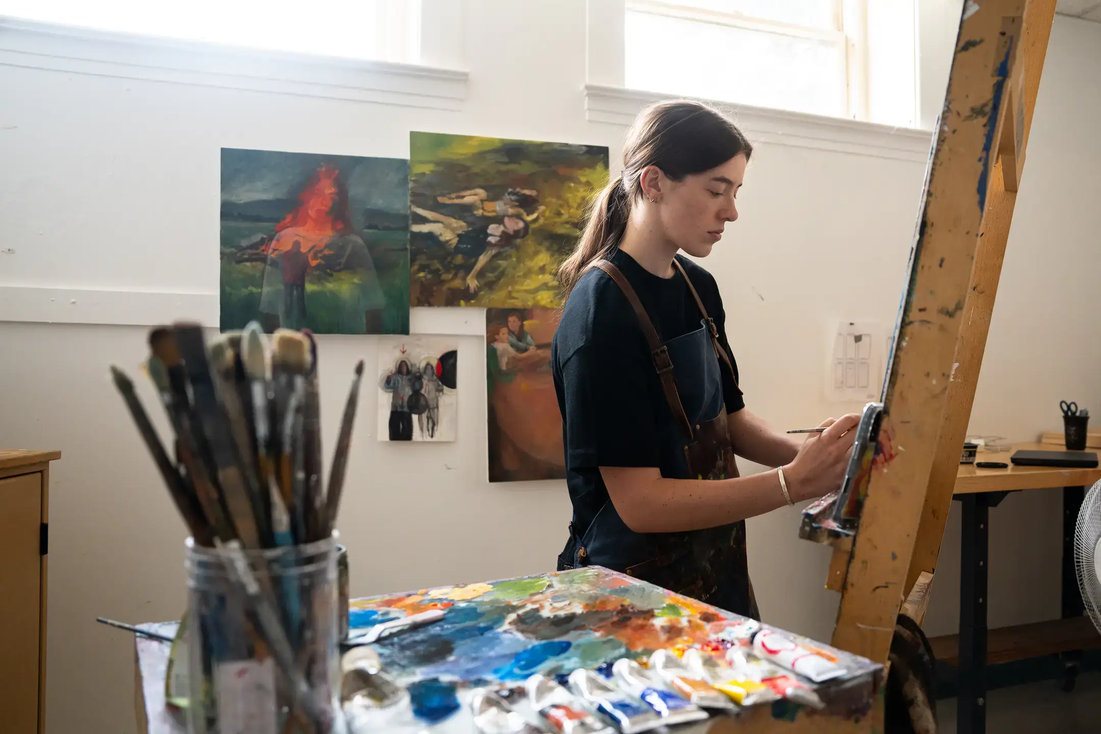 Woman in black shirt and blue overalls painting at her easel with paintings on the wall behind her and brushes in the foreground.