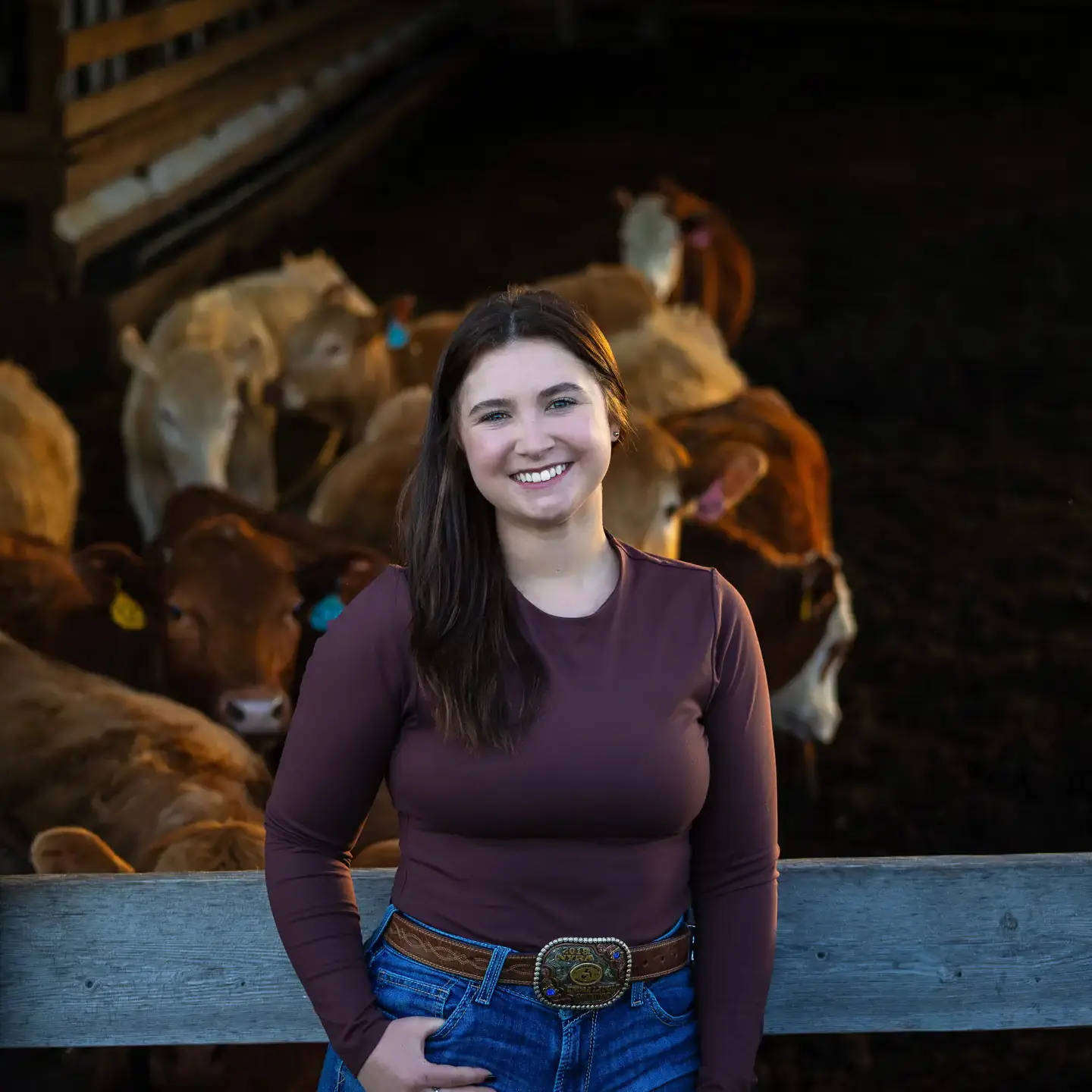 Smiling woman in jeans stands by a fence with cattle gathered inside a dim barn behind her