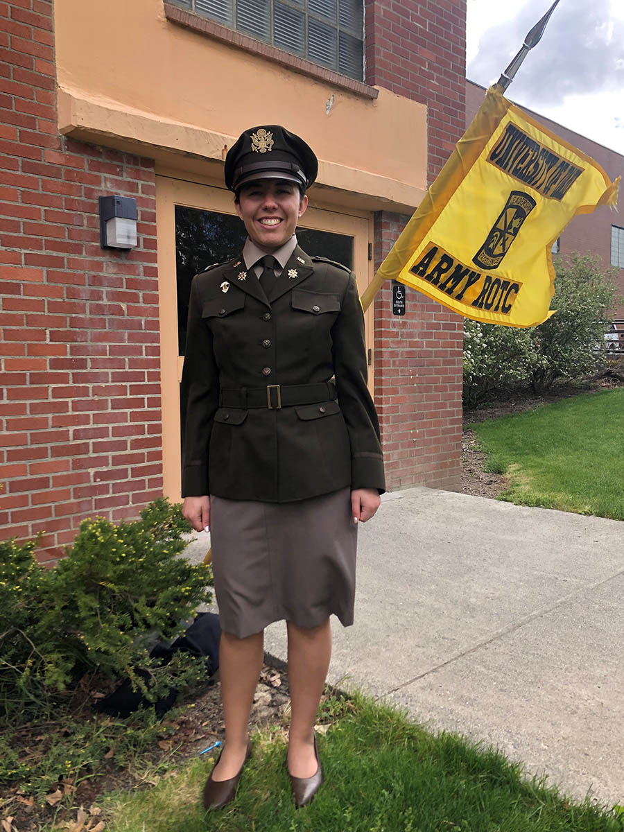 Woman in Army green dress uniform stands in front of ROTC guidon.
