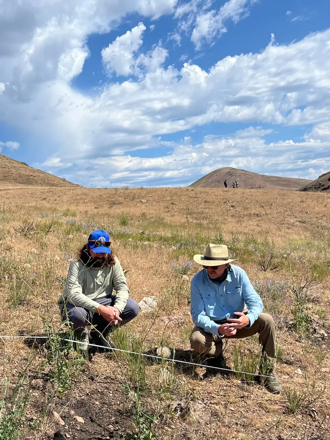 Two men wearing sunglasses and hats squat low to the ground to look at a patch of vegetation.