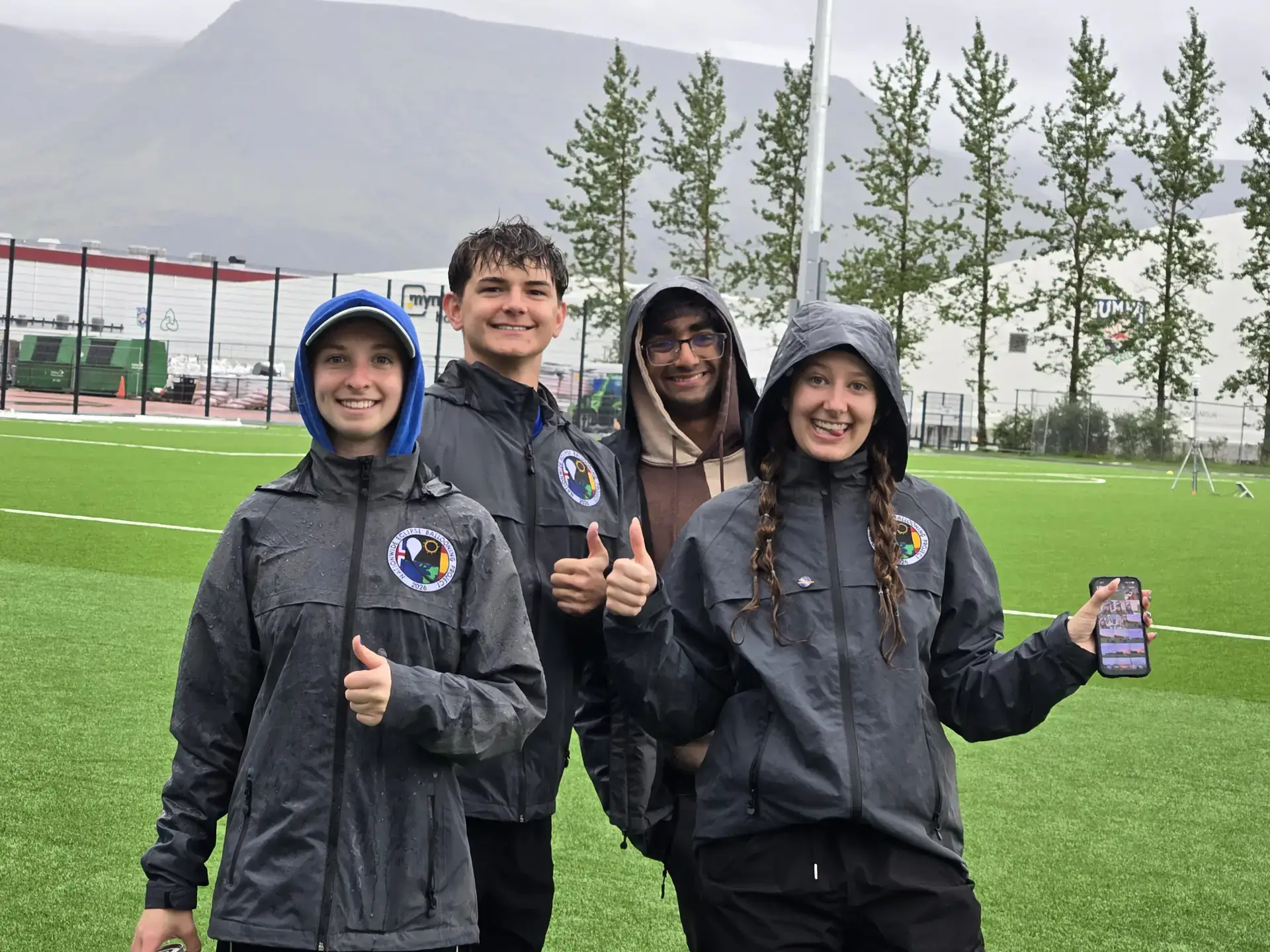 Four smiling people in rain jackets give thumbs up on a wet field, with mountains and trees behind them.