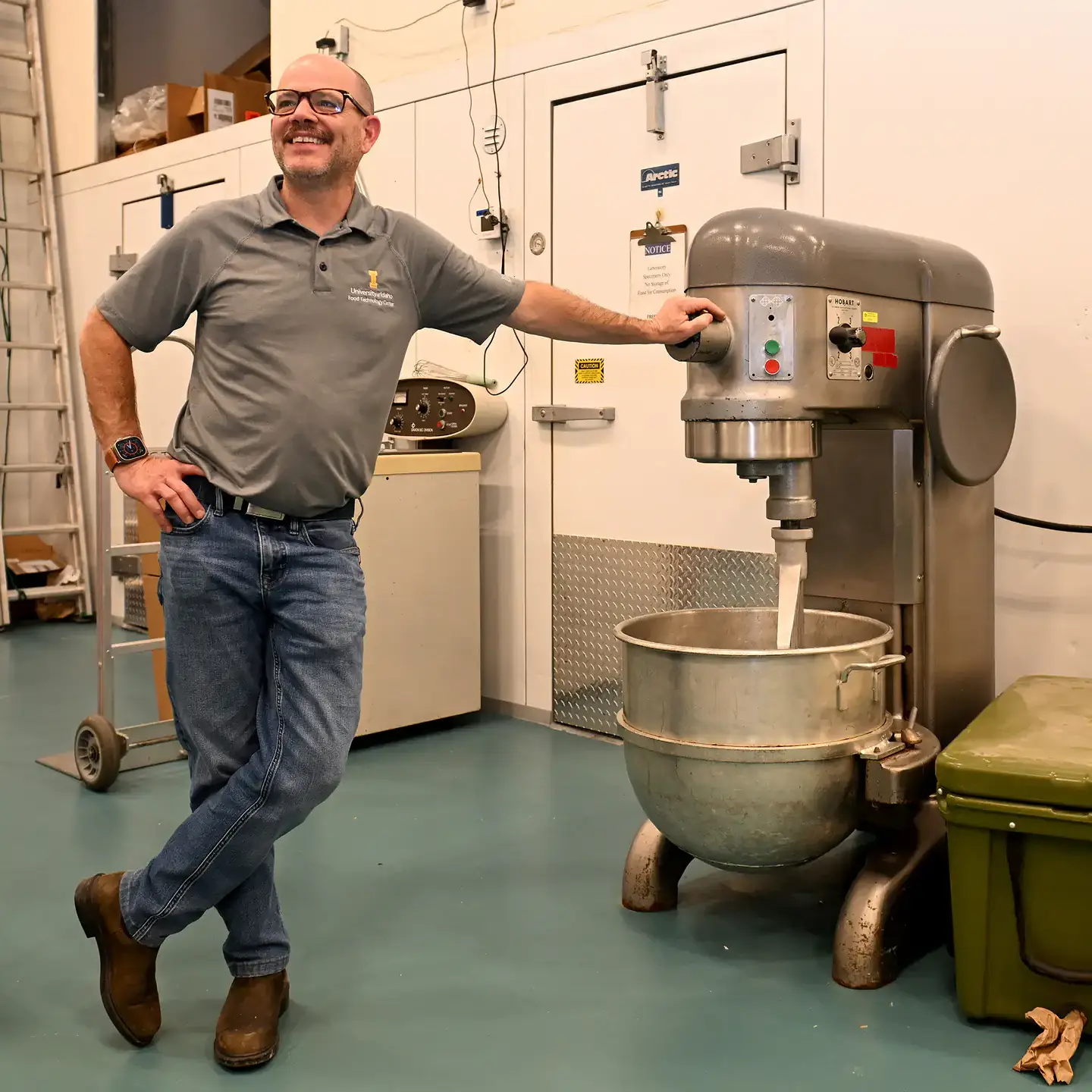 A person standing next to an industrial mixing machine in a workshop or lab, smiling and wearing a gray shirt and jeans.