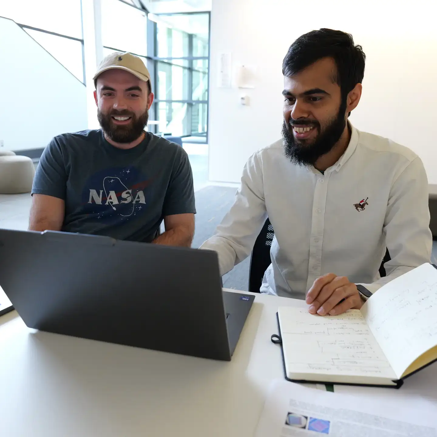 Two men collaborate at laptops with a notebook open on a desk in a bright office.