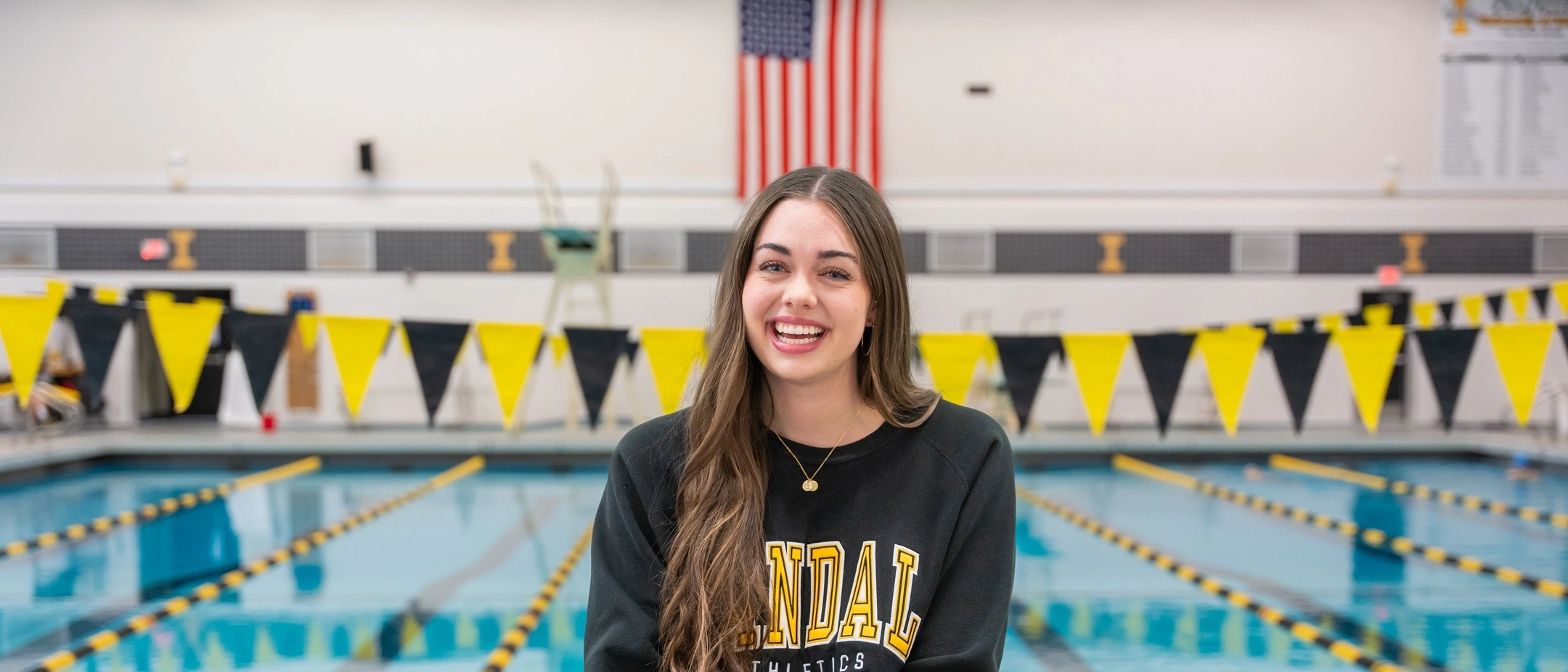 Smiling woman wearing a vandal sweatshirt sits on a diving platform above a college swimming pool.