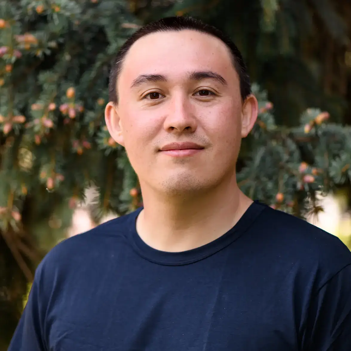 A person wearing a navy shirt, standing outdoors near a pine tree, looking at the camera with a slight smile.