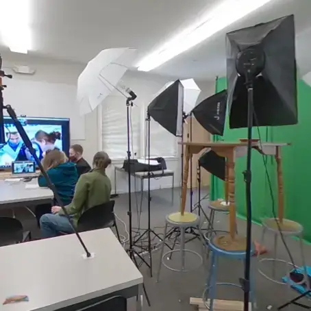 Students in room with a green screen, lighting equipment, and a large screen.