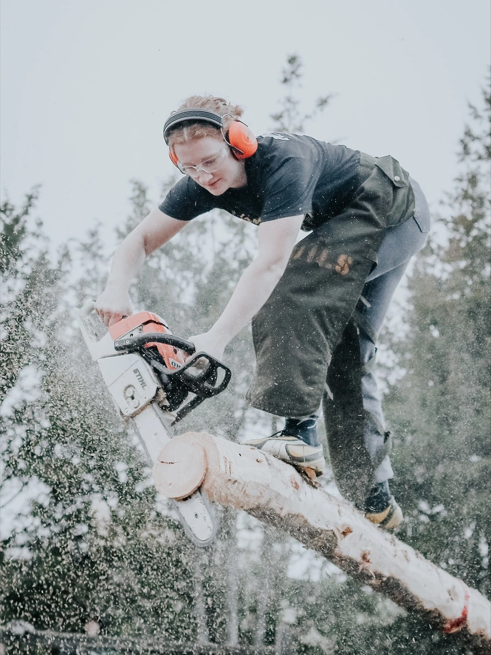 Woman stands on an elevated log cutting the end with a chainsaw.