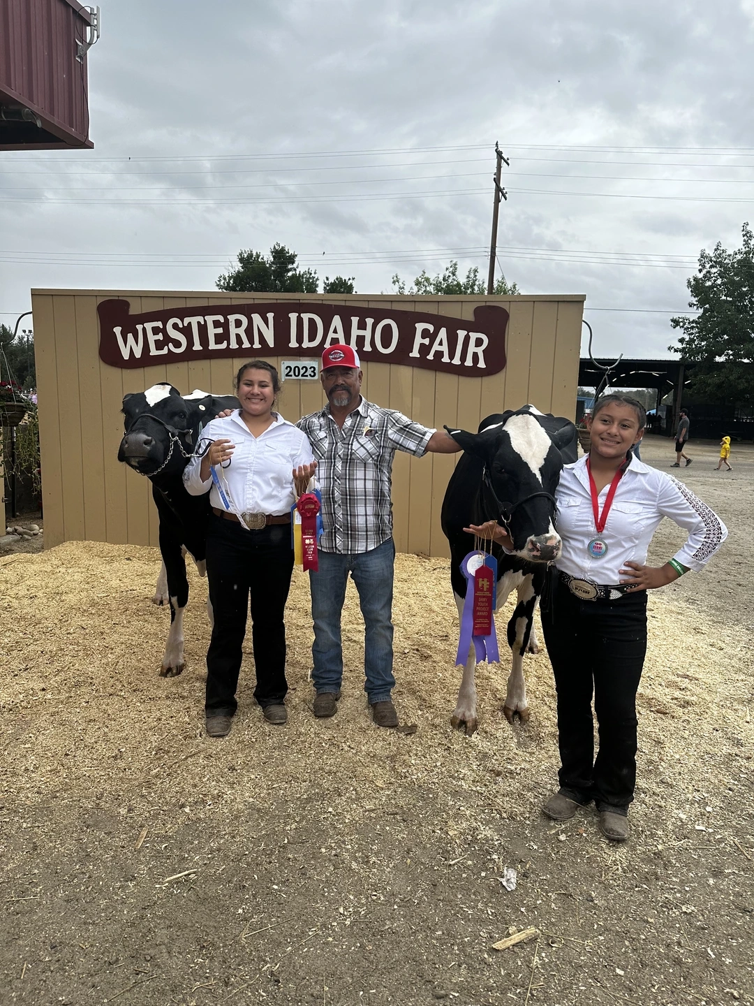 Two girls and their grandfather pose with dairy heifers and blue ribbons in front of a sign that reads Western Idaho Fair. 
