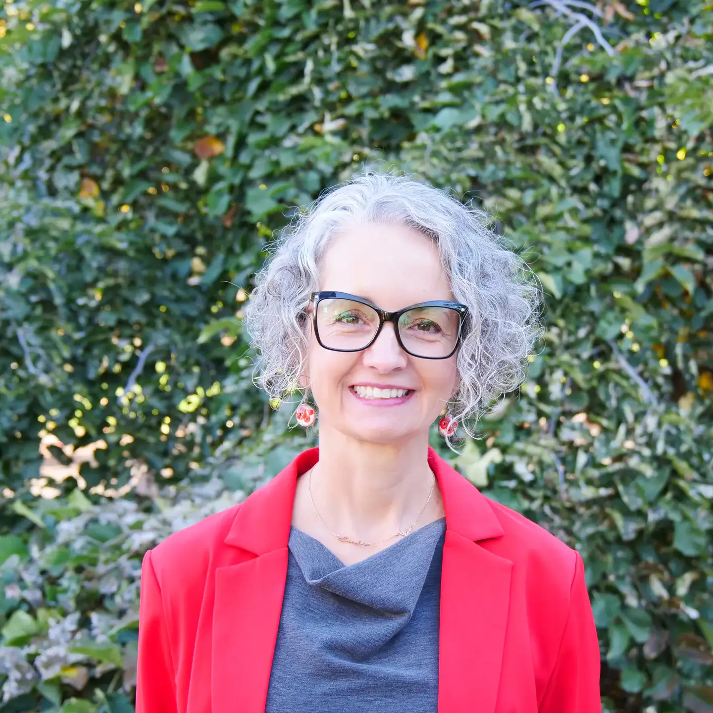 An individual with curly gray hair, wearing glasses, a red jacket, and a gray top, standing outdoors in front of greenery.
