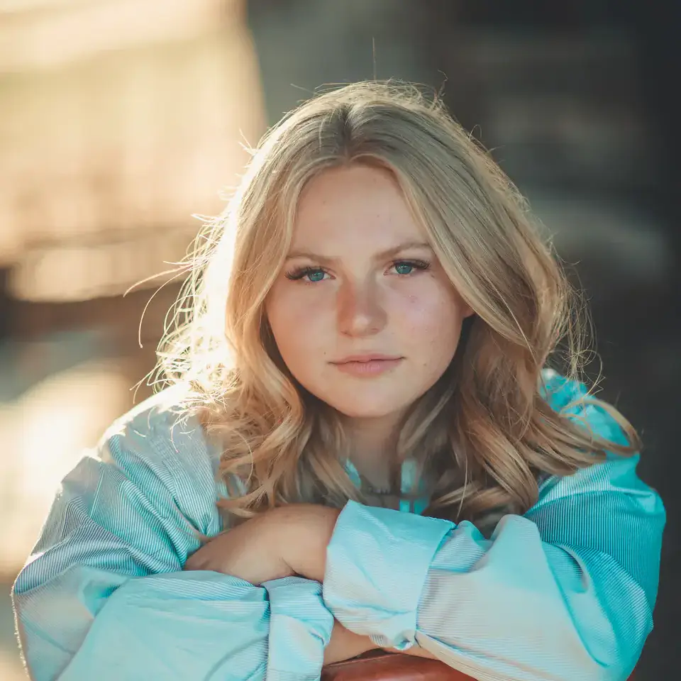 Portrait of a blonde young woman in a light blue blouse, resting her arms on a chair outdoors