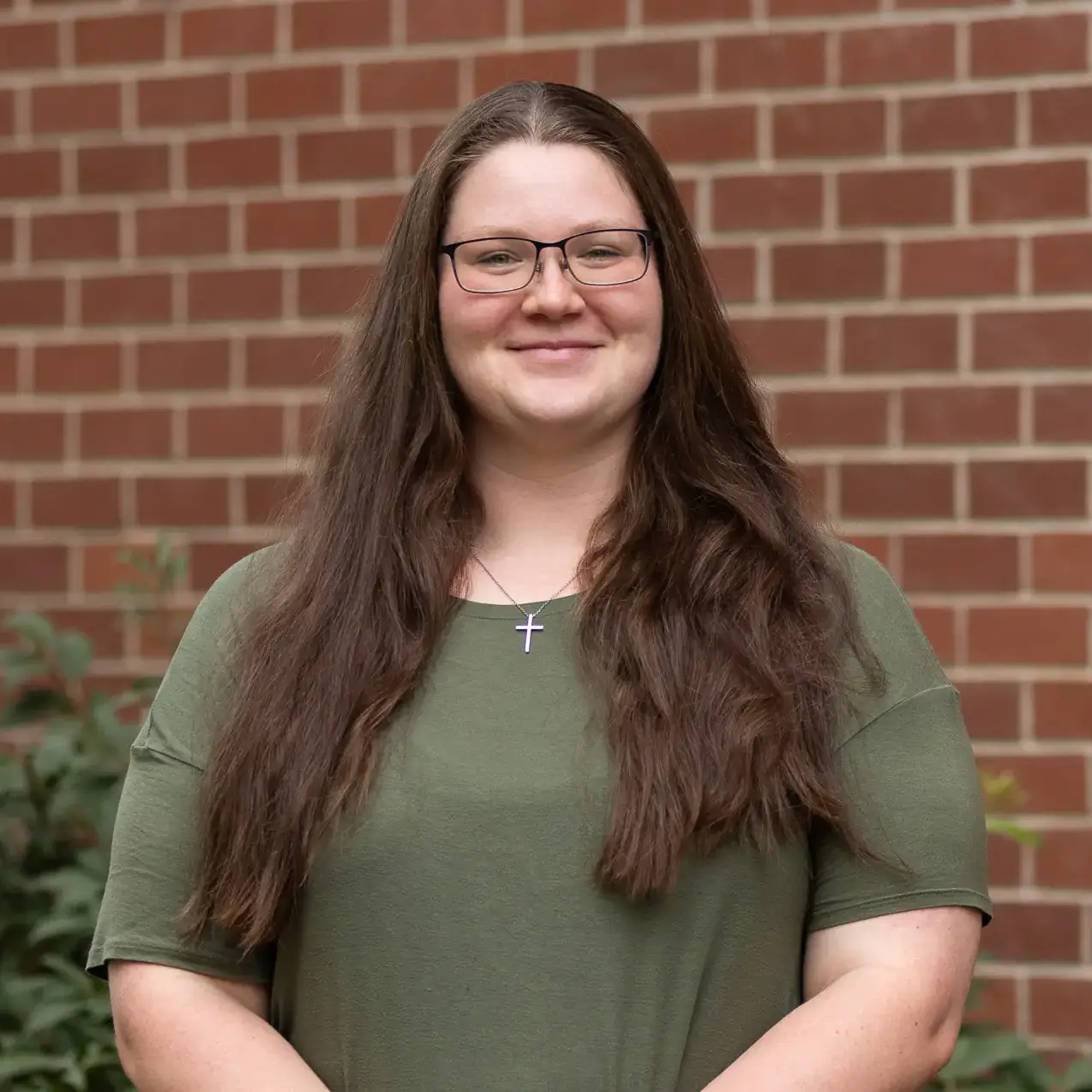 Smiling woman with long brown hair and glasses standing in front of a brick wall.