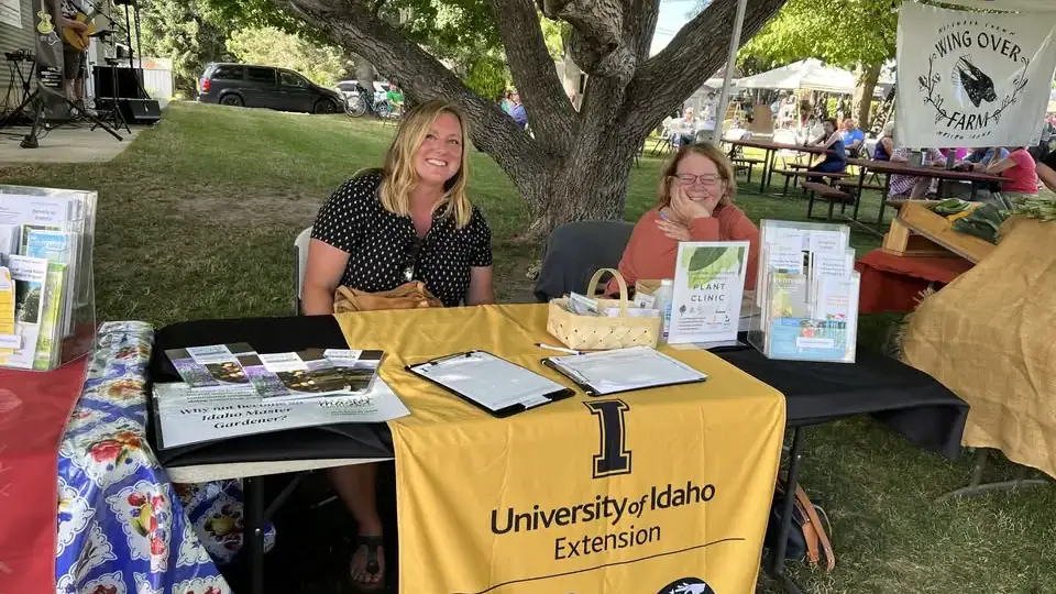 Two women sit at a booth with a University of Idaho Extension table cloth on a shady lawn.