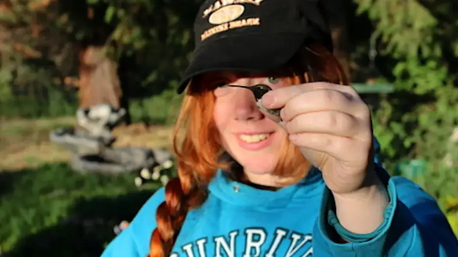 A woman wearing a hat outdoors gently holds a hummingbird keeping the wings folded to its body