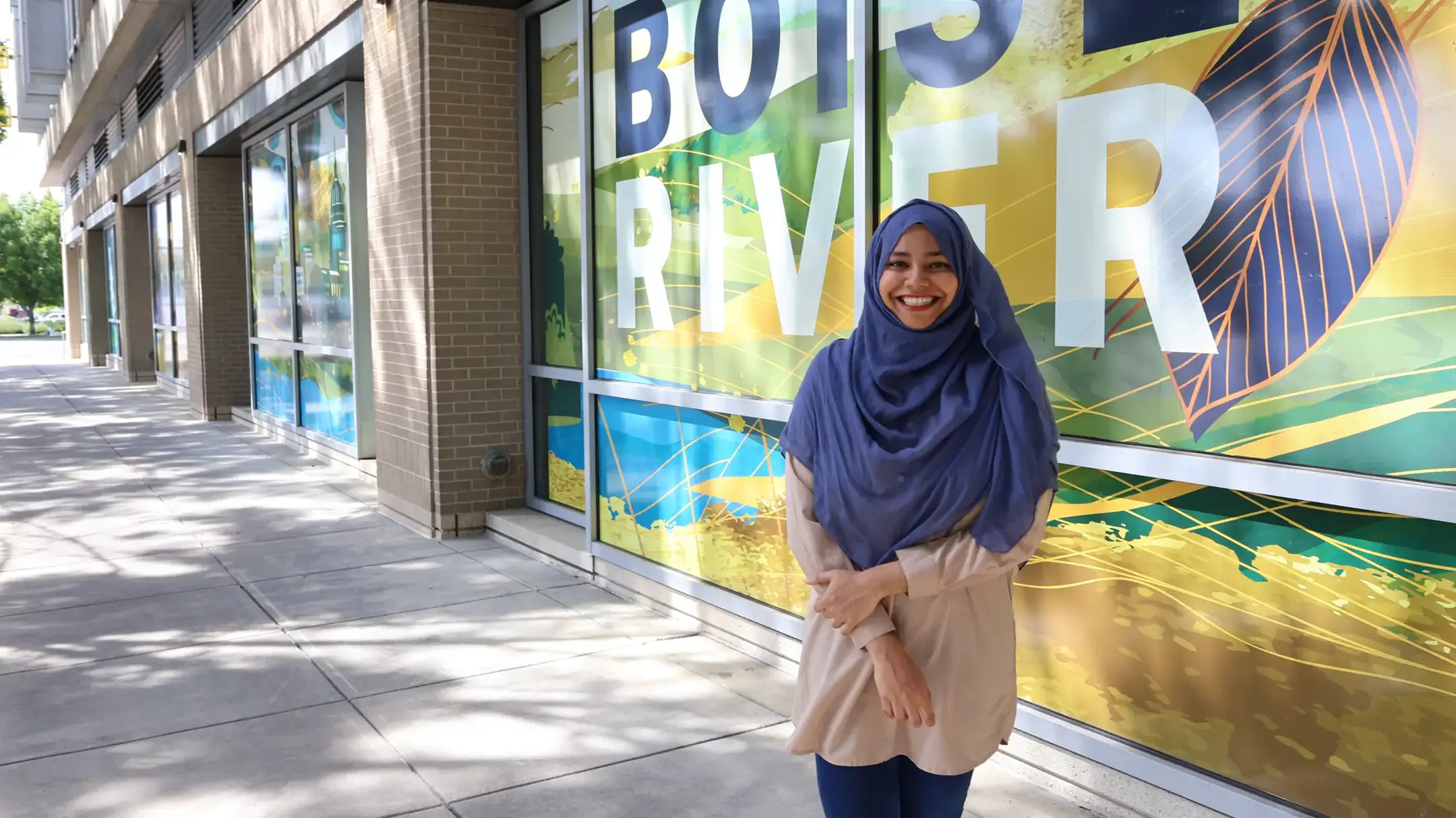 A woman standing and smiling outdoors, near colorful window signage reading 'Boise River' on a sunny day.