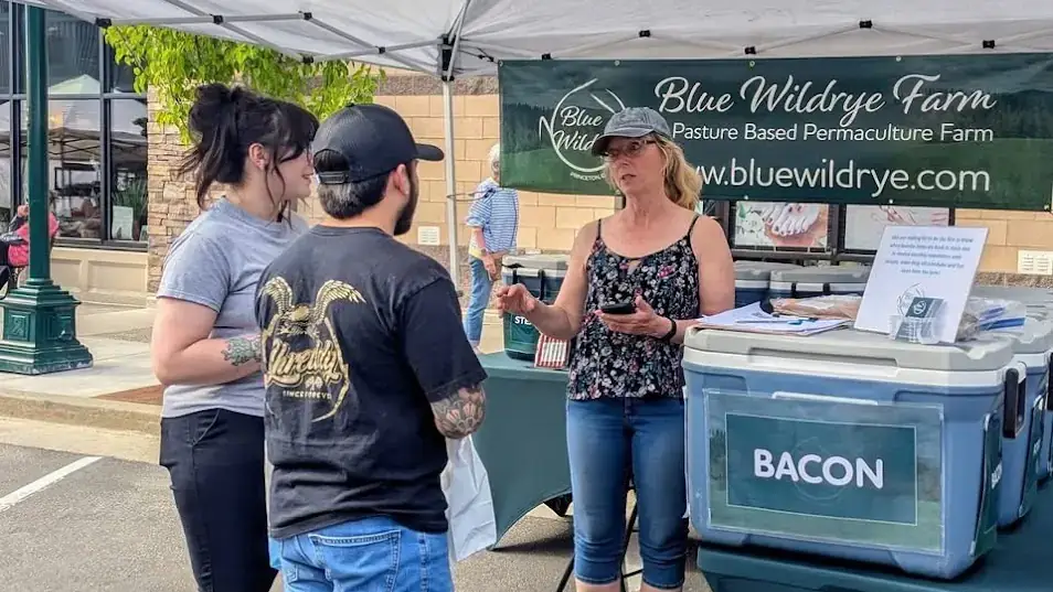 A woman standing under an awning talks with two customers, standing by a cooler filled with bacon.