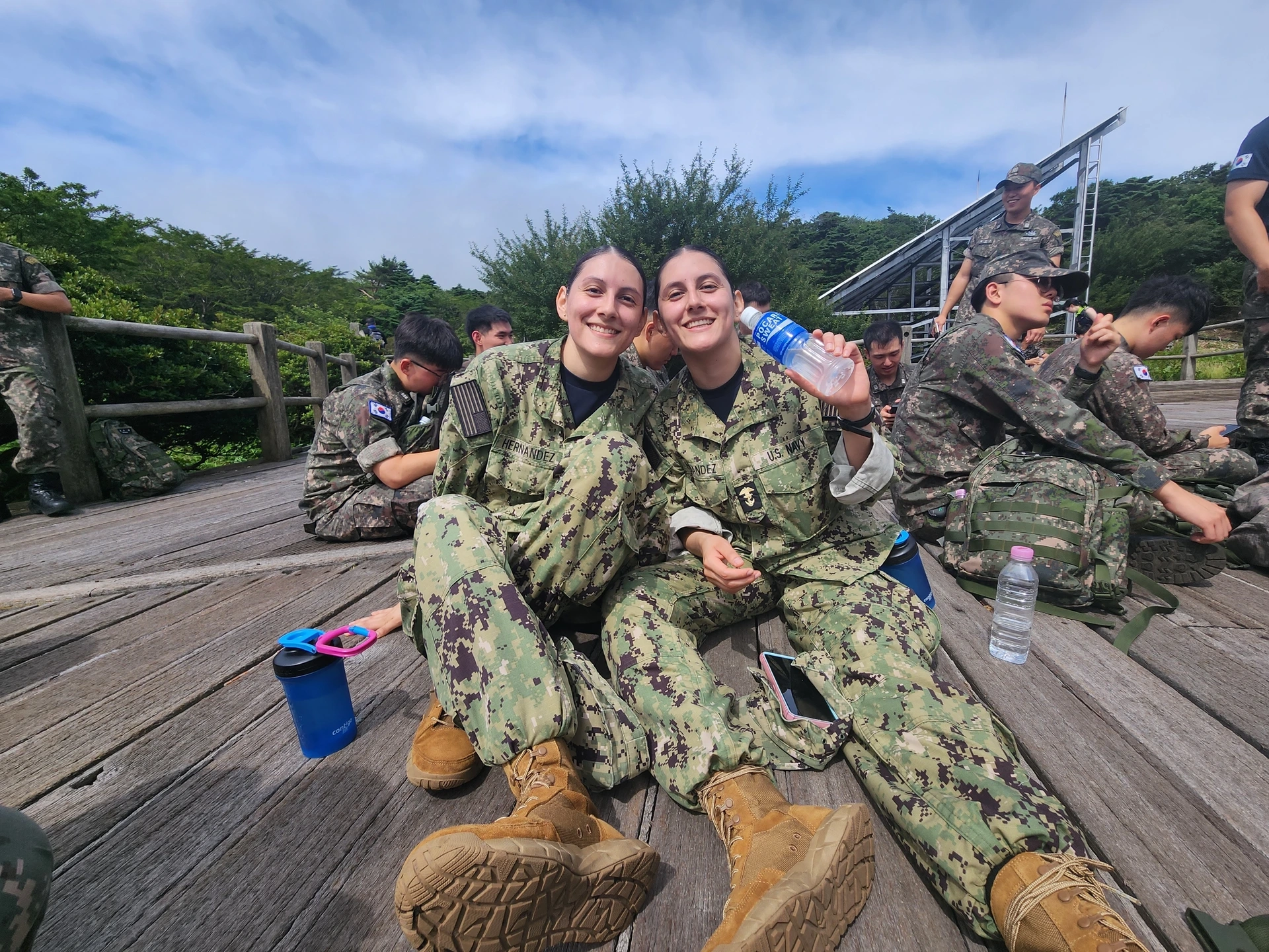 Two women wearing BDUs hug while sitting on a wooden deck.