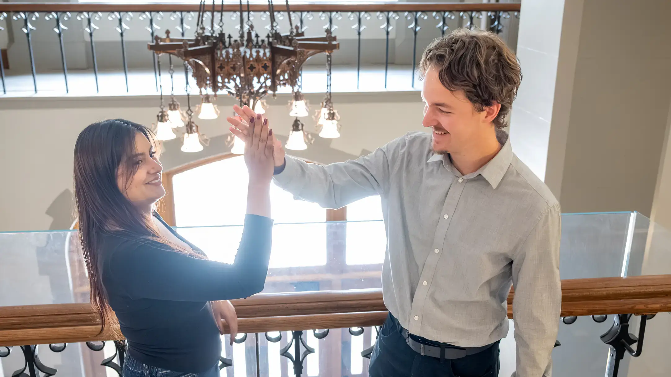 Two people high-fiving while smiling in a brightly lit interior with a chandelier visible in the background.