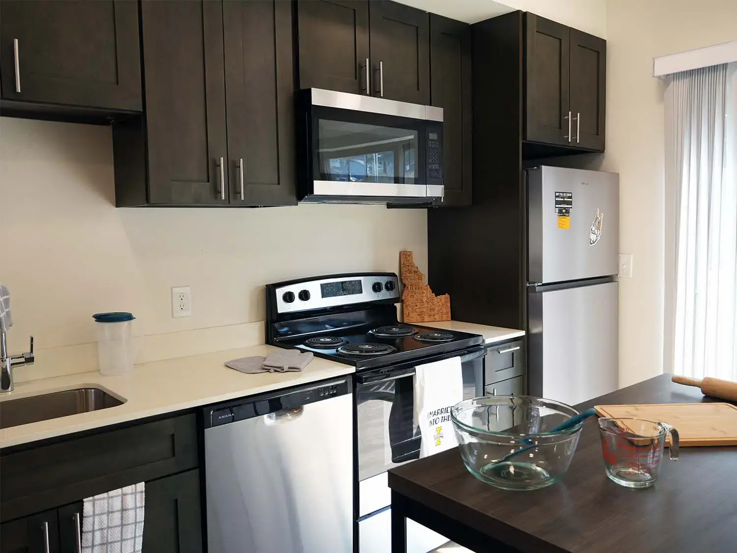 Modern kitchen in a Vandal Village Family Housing apartment with dark cabinetry, stainless steel appliances, white countertops, and an island workspace.