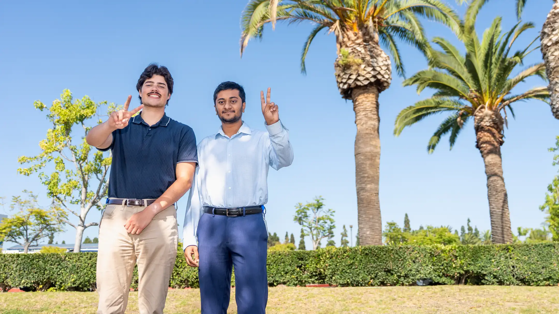 Two individuals stand outdoors under palm trees, smiling and gesturing towards the camera, with greenery and clear blue sky in the background.