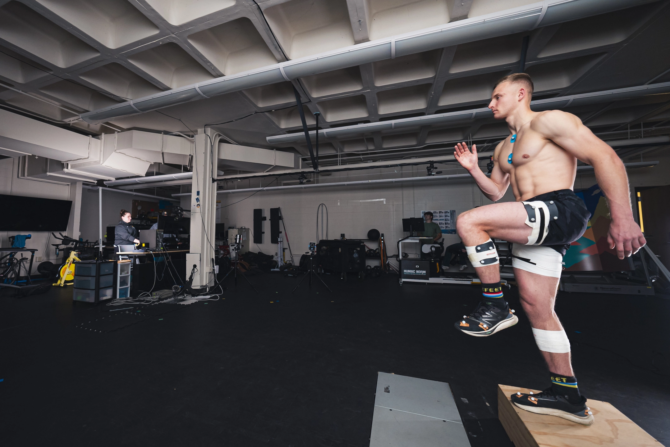 Shirtless man exercises by stepping on and off a wooden box to the right of frame, while a woman works at a computer on the left side.