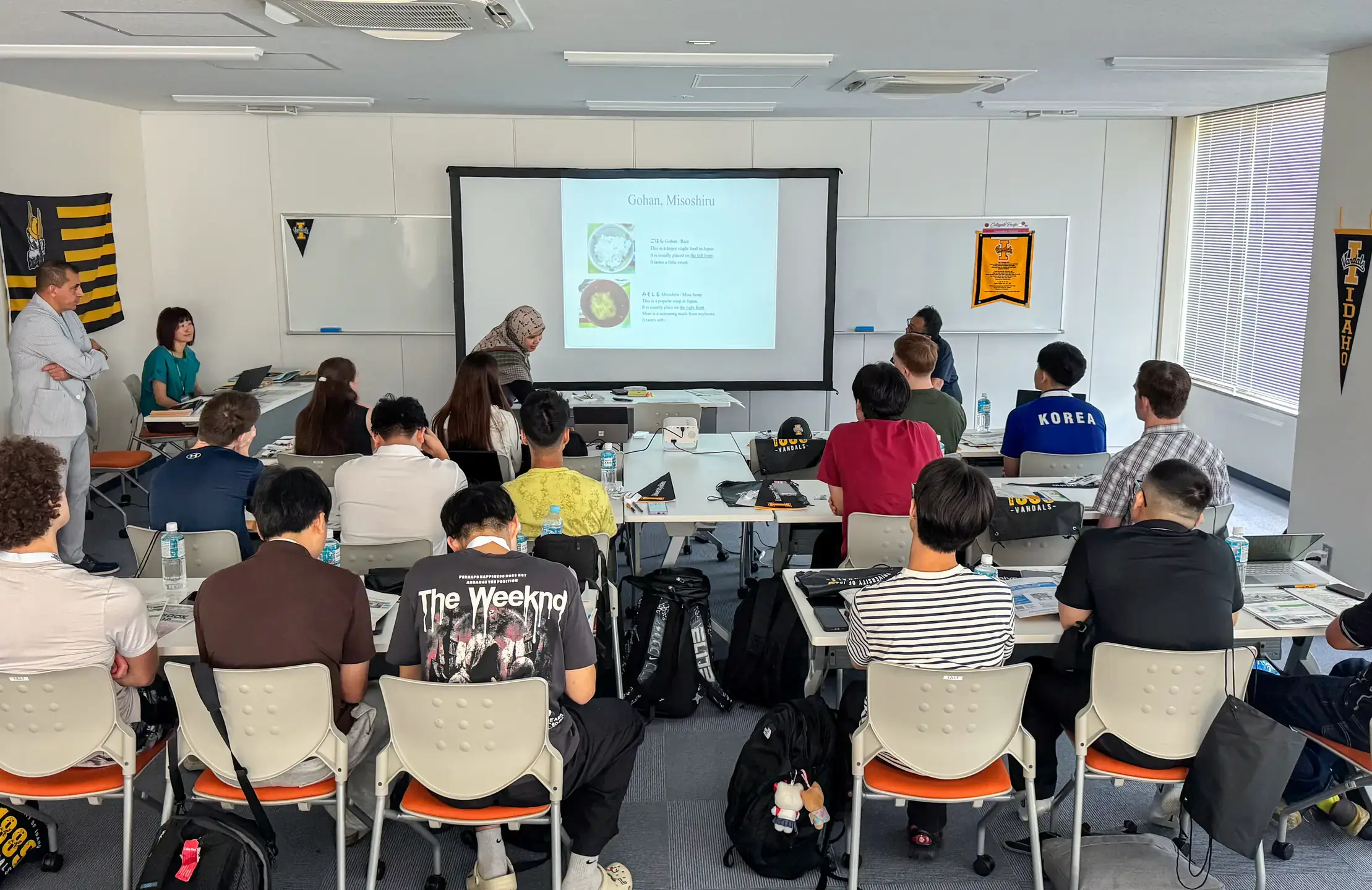 Classroom setting with individuals seated at tables facing a presentation screen displaying a slide about Japanese cuisine.
