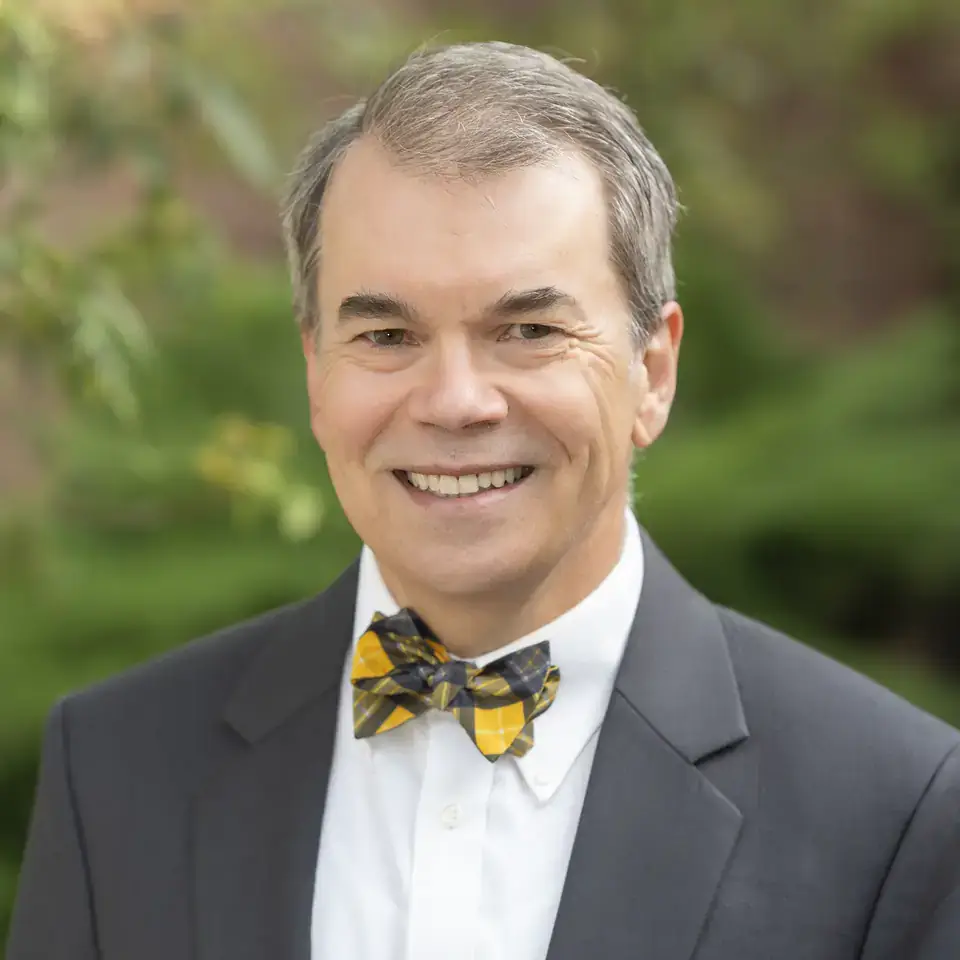 A person wearing a suit and a patterned bow tie, smiling in an outdoor setting with greenery in the background.
