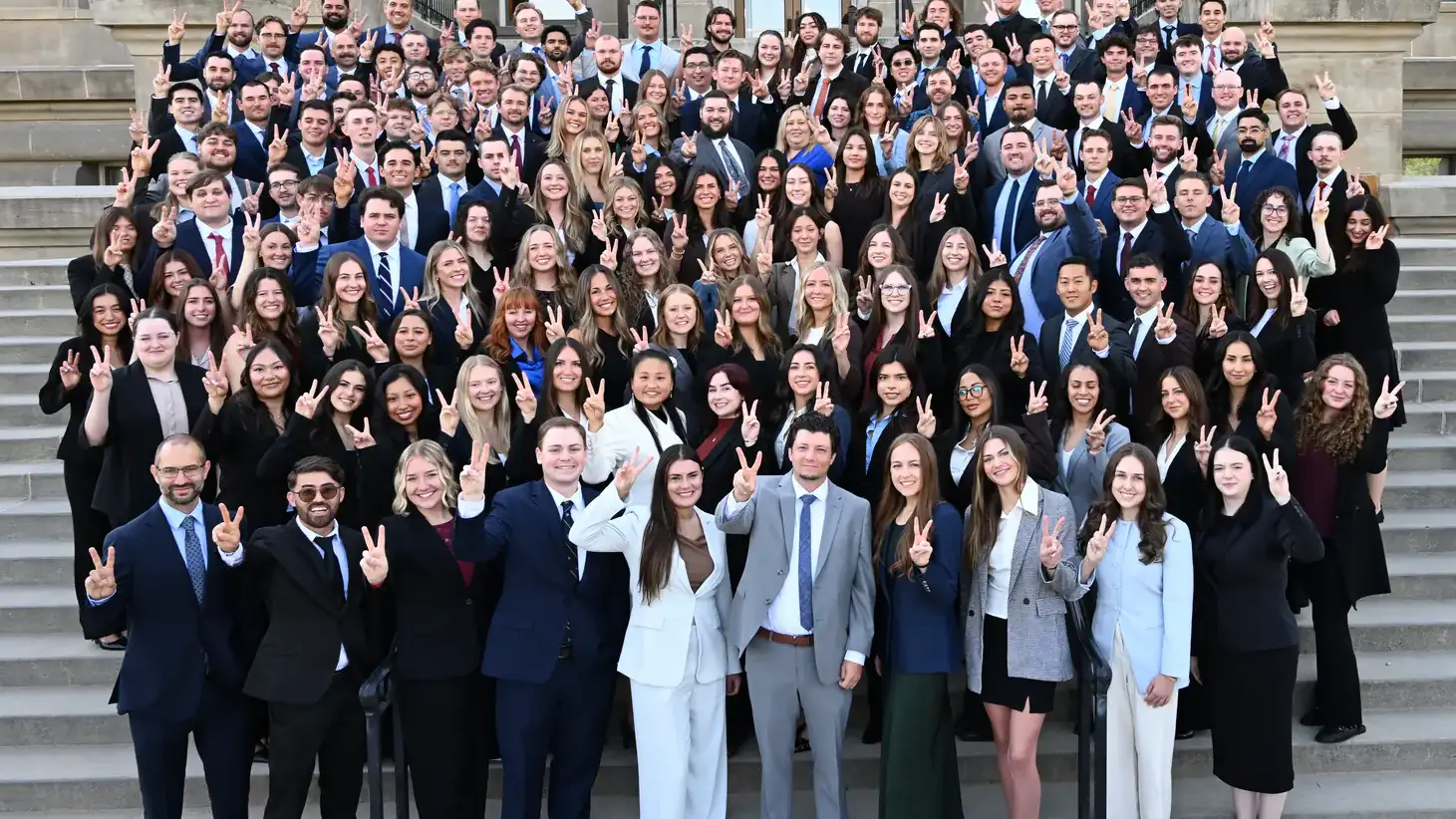 A large group of people posing on stairs in front of a stone building, many making hand gestures with peace signs.
