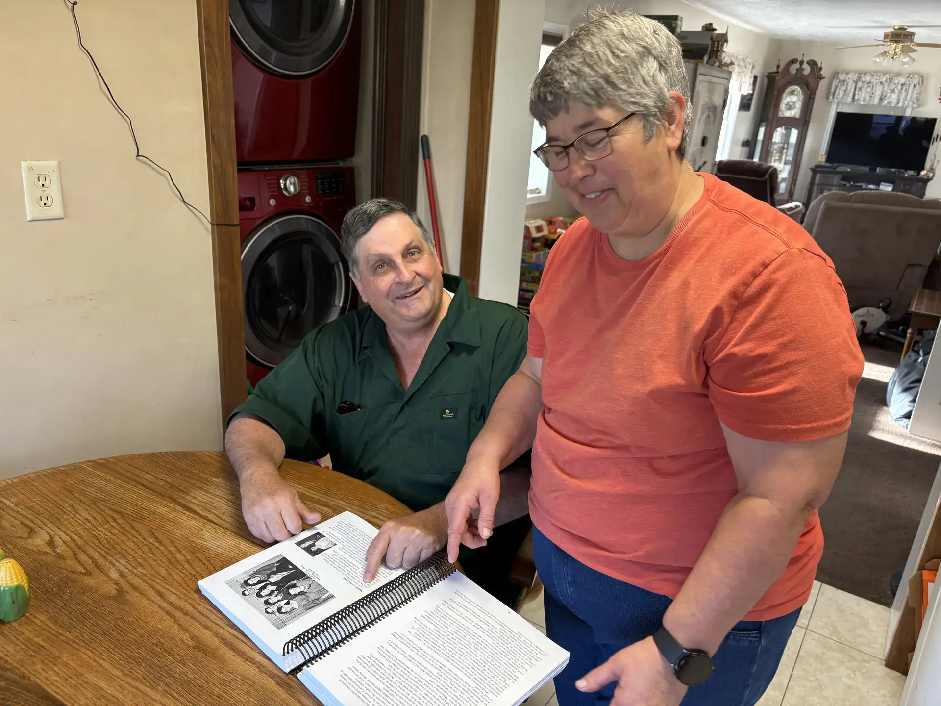 Two people look at a book on a kitchen table.