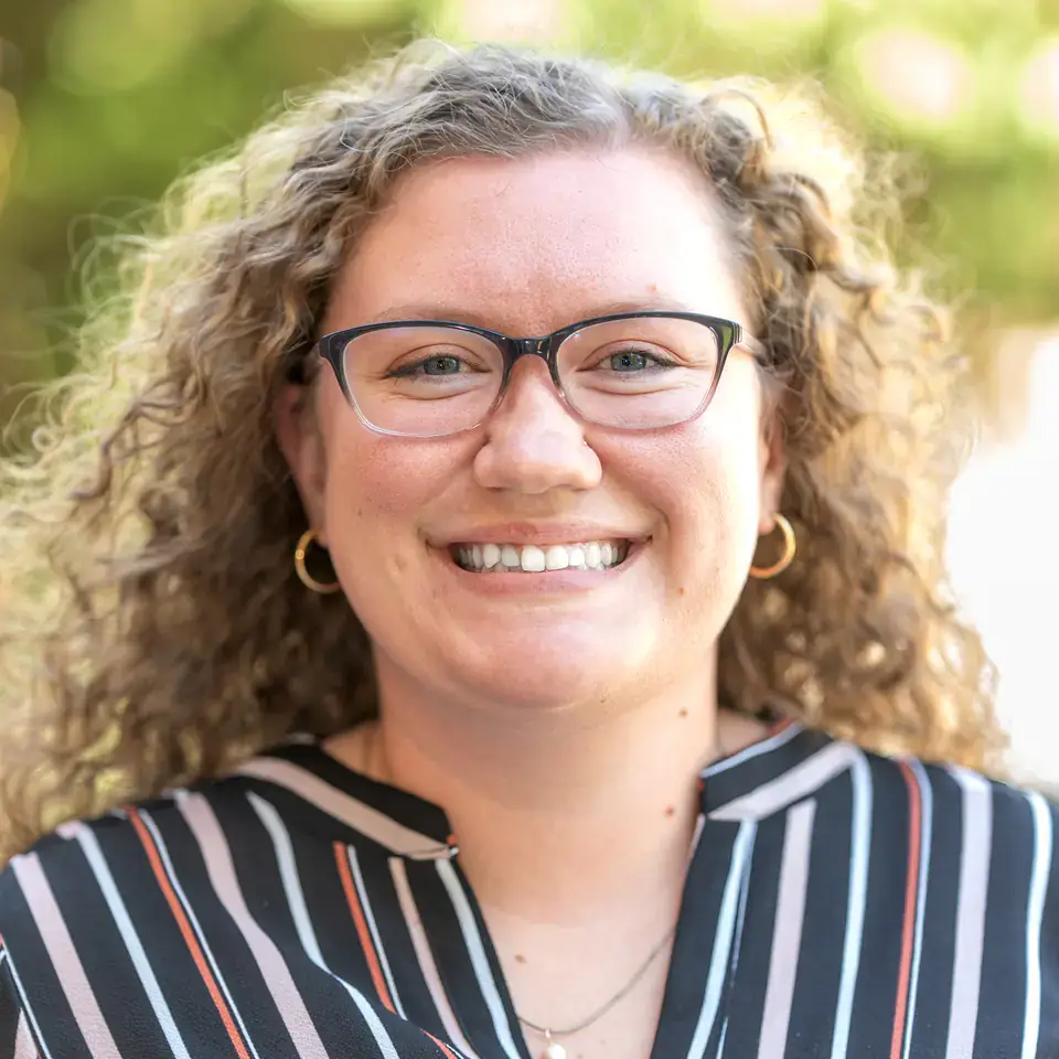A person with curly hair and glasses is smiling while wearing a striped shirt with a natural background.