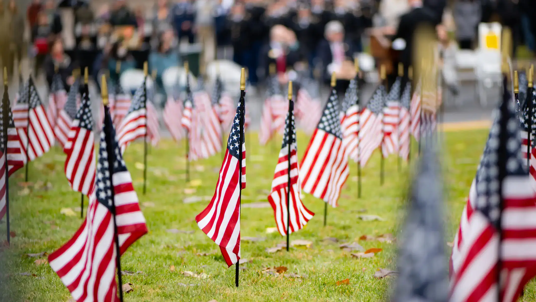 Flags are placed in a grid, rising from Admin Lawn, with a crowd of out of focus memorial attendees in the background.