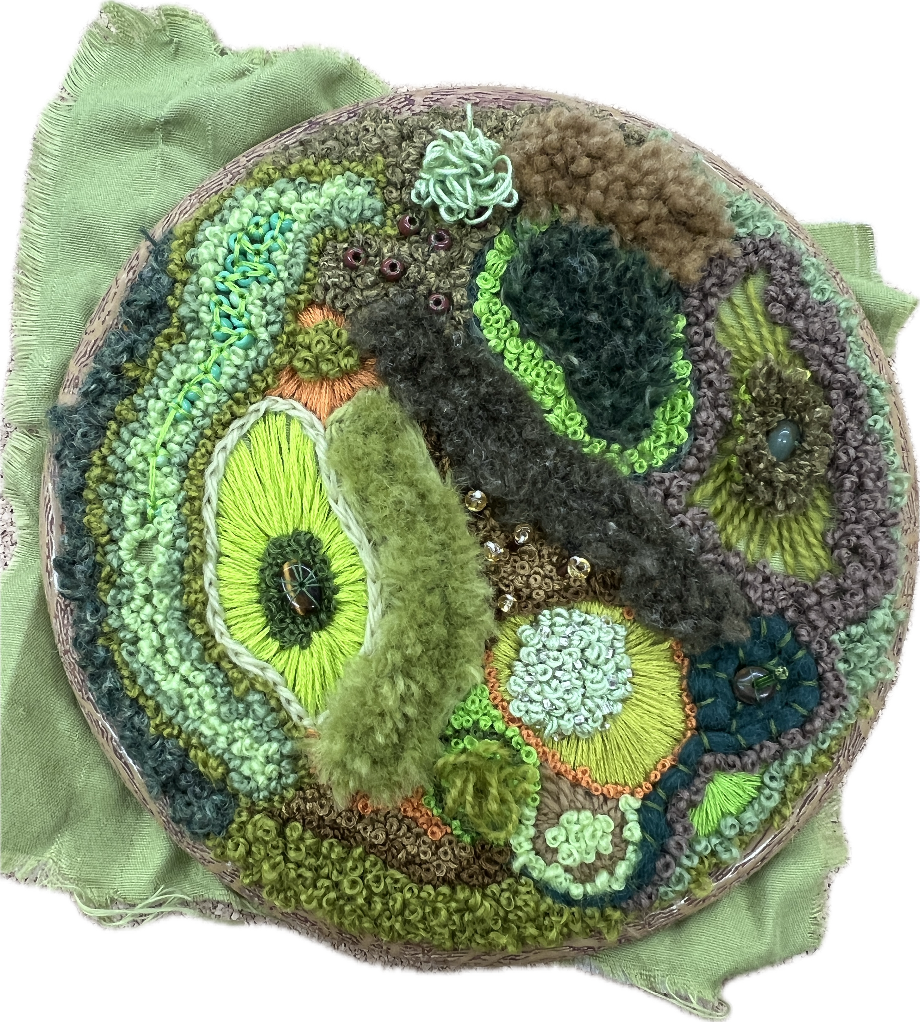 A circular embroidery featuring moss-inspired textures and colors, with greens, browns, and blue shades creating an organic pattern.