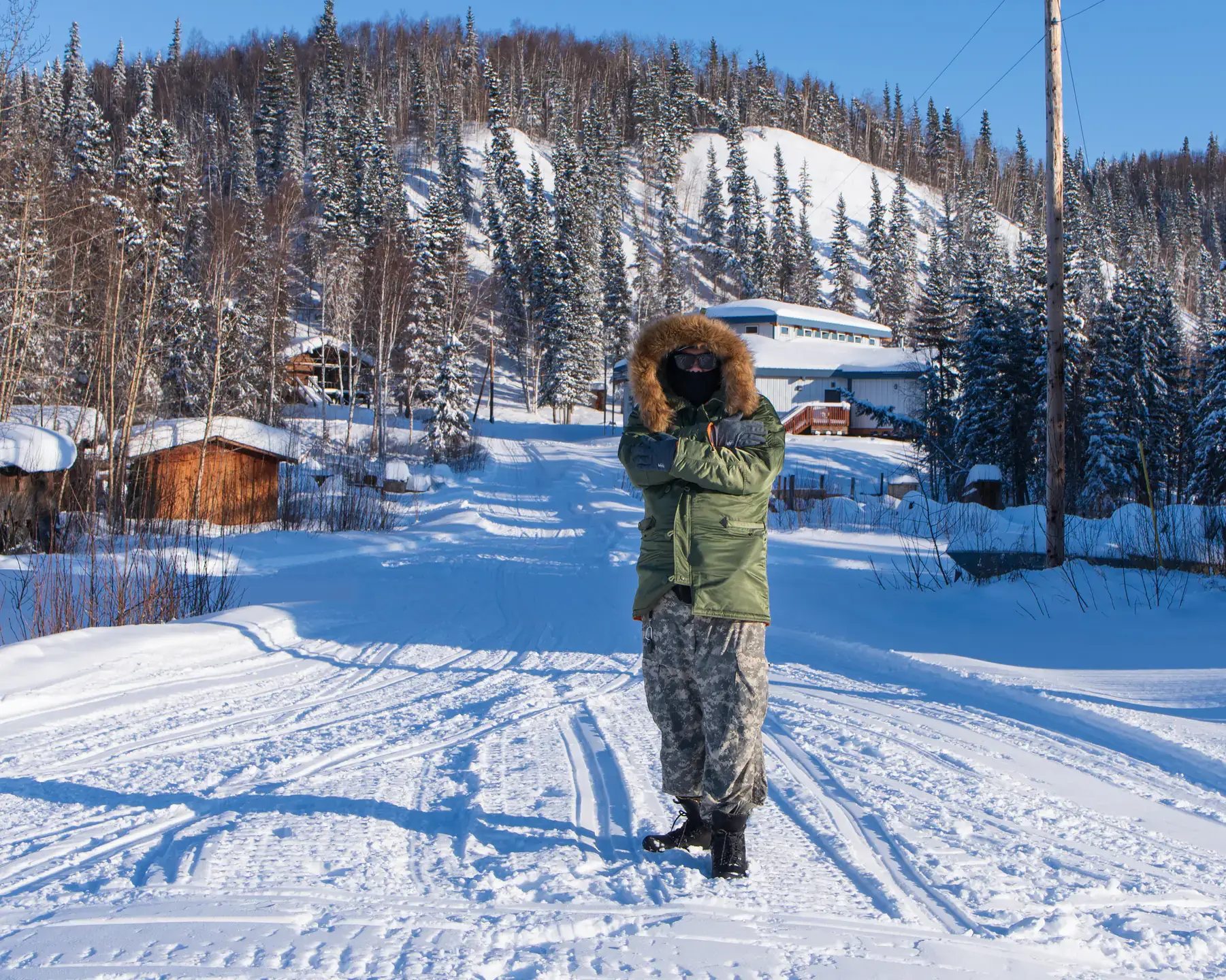 Young Sim, wearing a heavy winter coat, stands on snow-covered street with several small buildings, with a mountain and pine trees in the background.