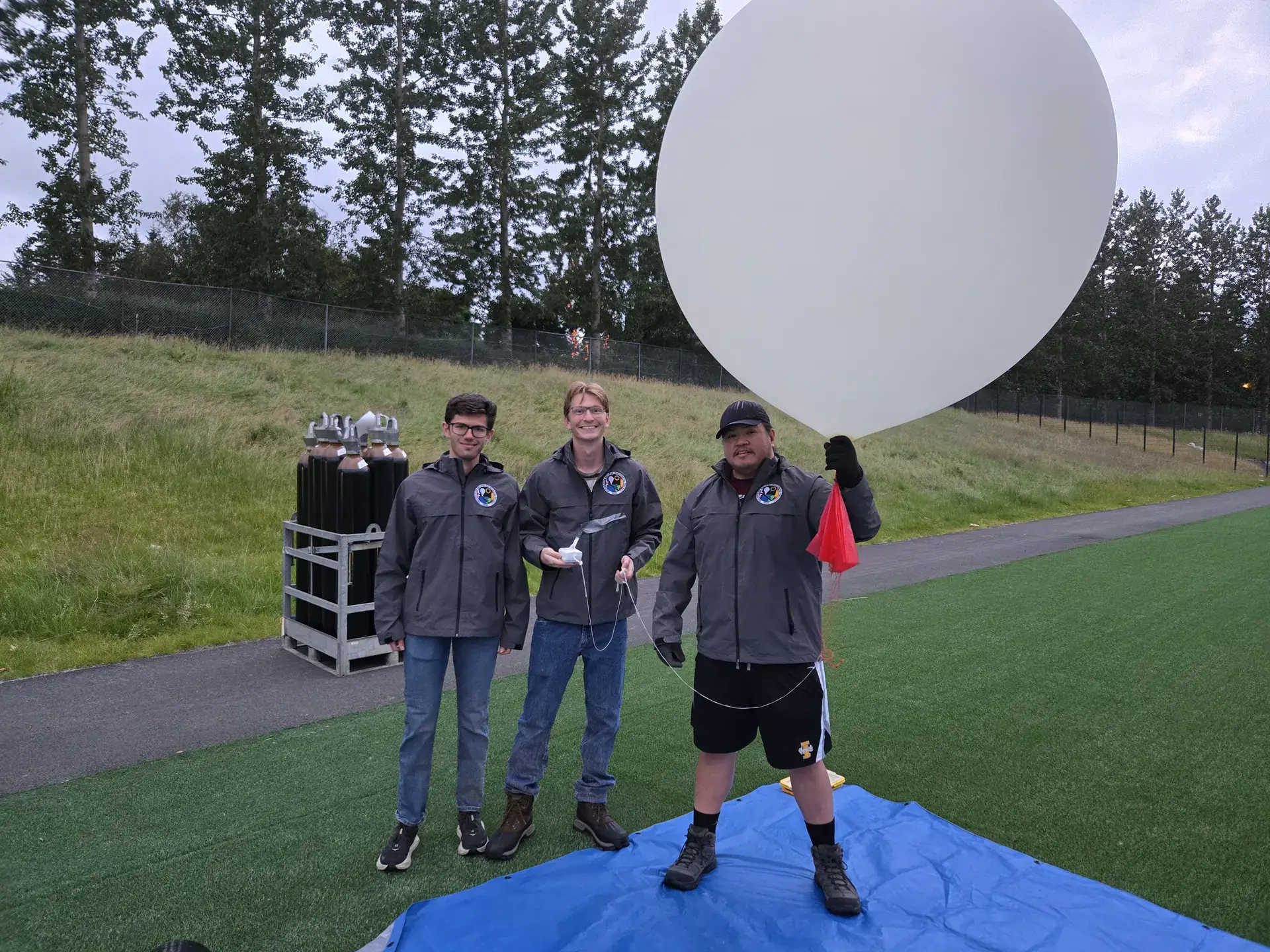 Three people in matching jackets stand on a field, preparing a large white weather balloon for launch.