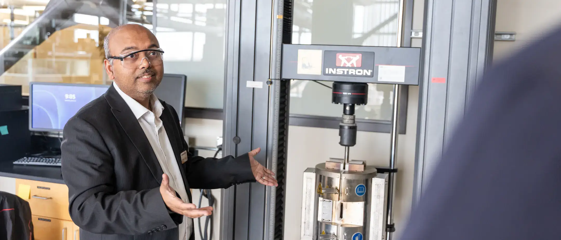 A person in formal attire gestures towards a mechanical device in a laboratory setting.