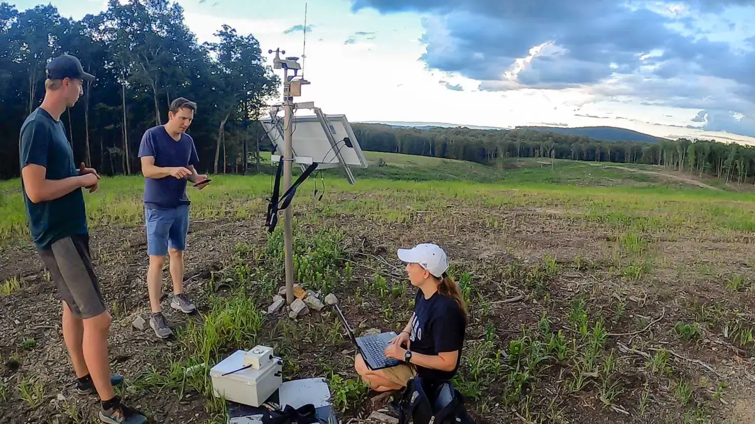 Three people stand on a hill in an open meadow near a miniature weather center surrounded by a forest. 