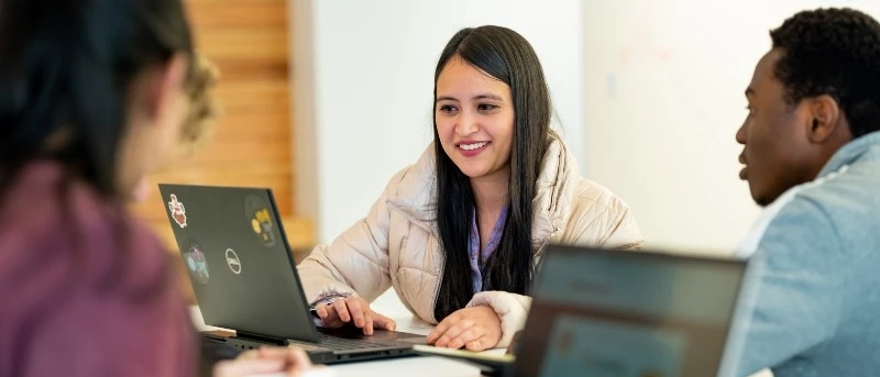 Three students smile and laugh while working on a group project together on their laptops. 