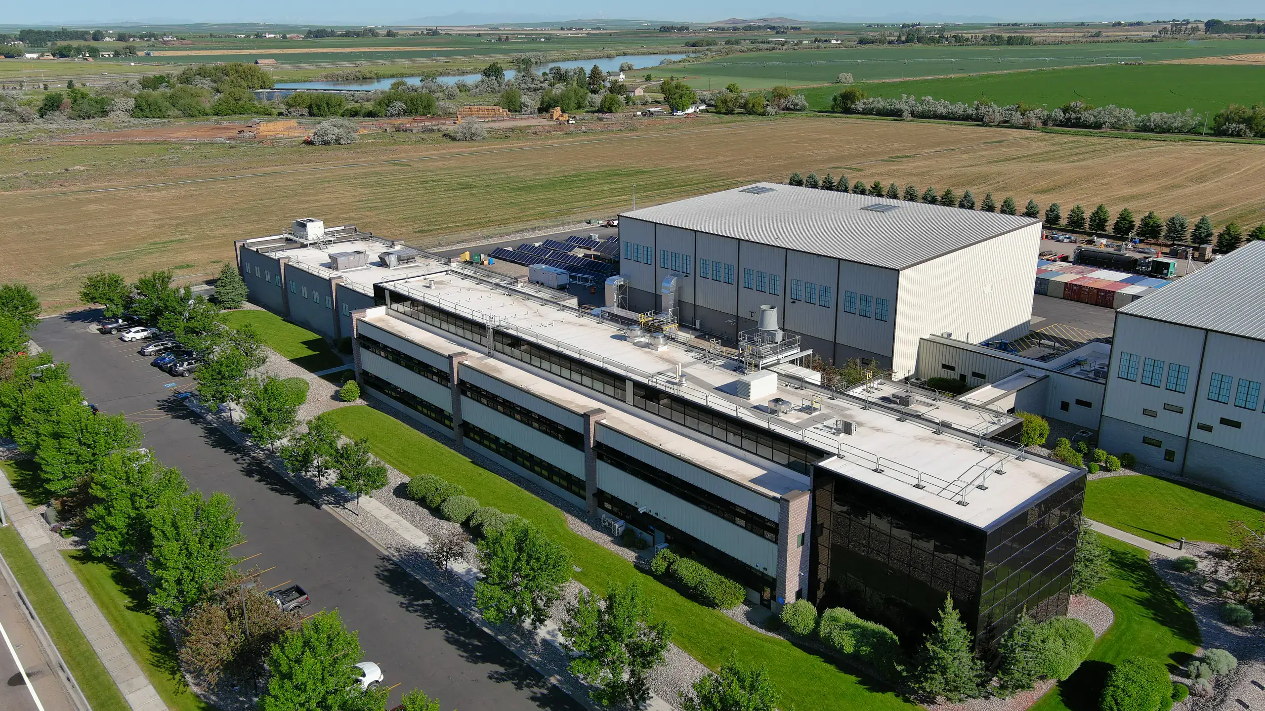 Aerial view of a large industrial research building beside parking lot, trees, and open farm fields.