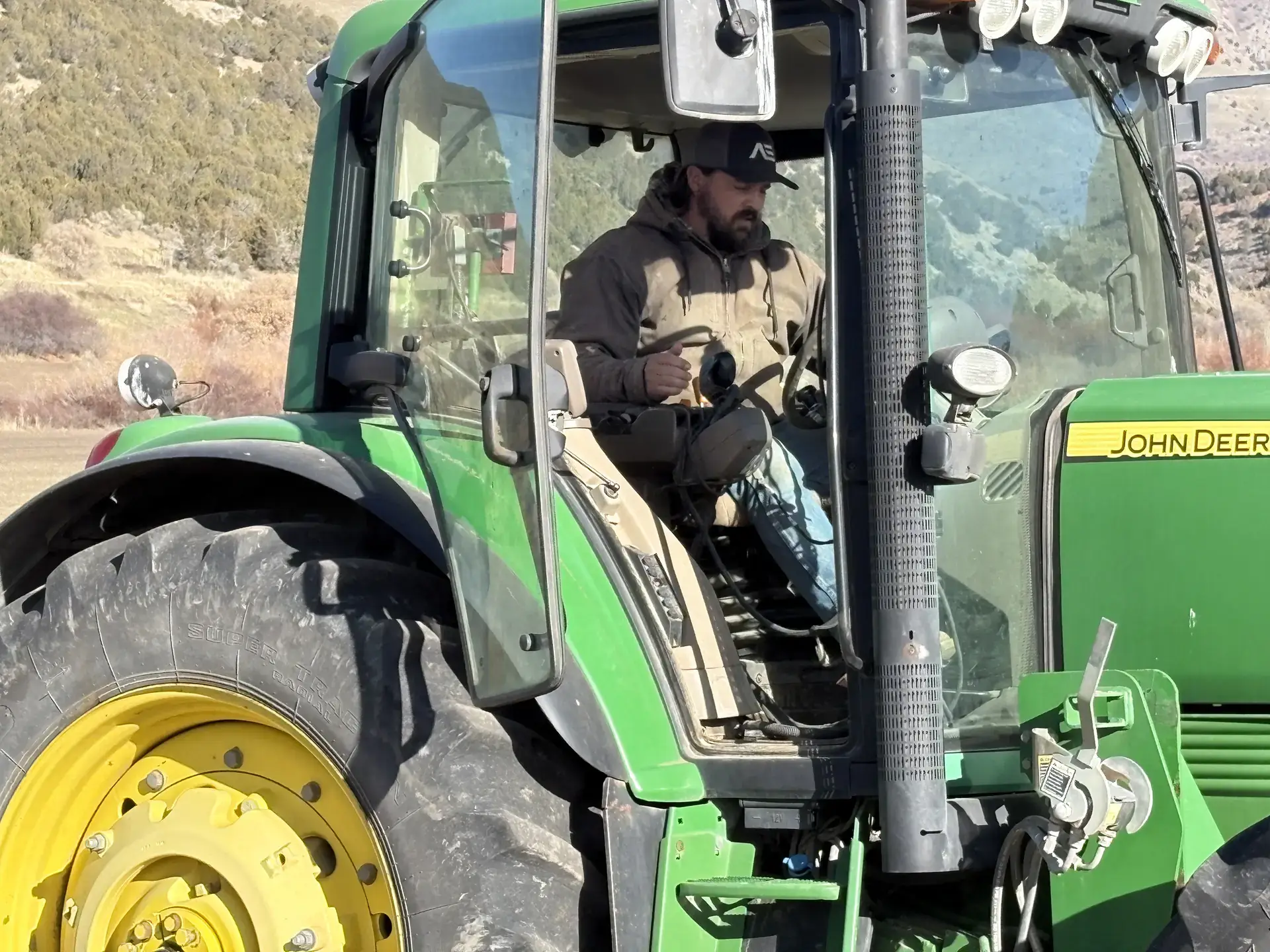 A young farmer sits in a tractor with an open door in the midst of a farm field. 