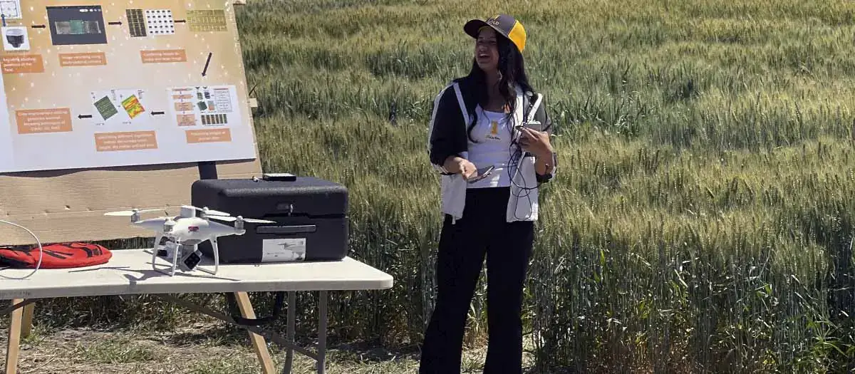 Pabitra Joshi stands in a field beside a table with a drone and equipment with a process poster on display in the background.