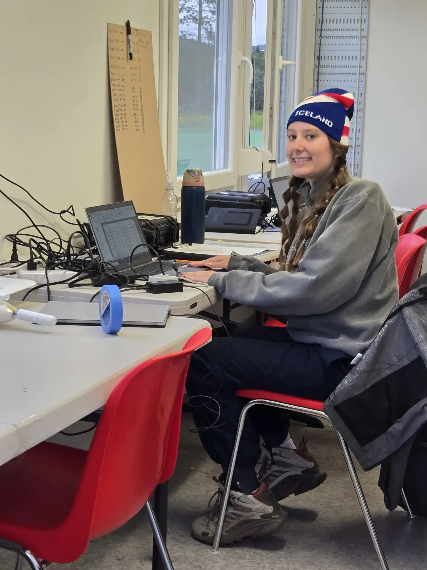 Person in an Iceland beanie smiling while working on a laptop at a table with cables and equipment.