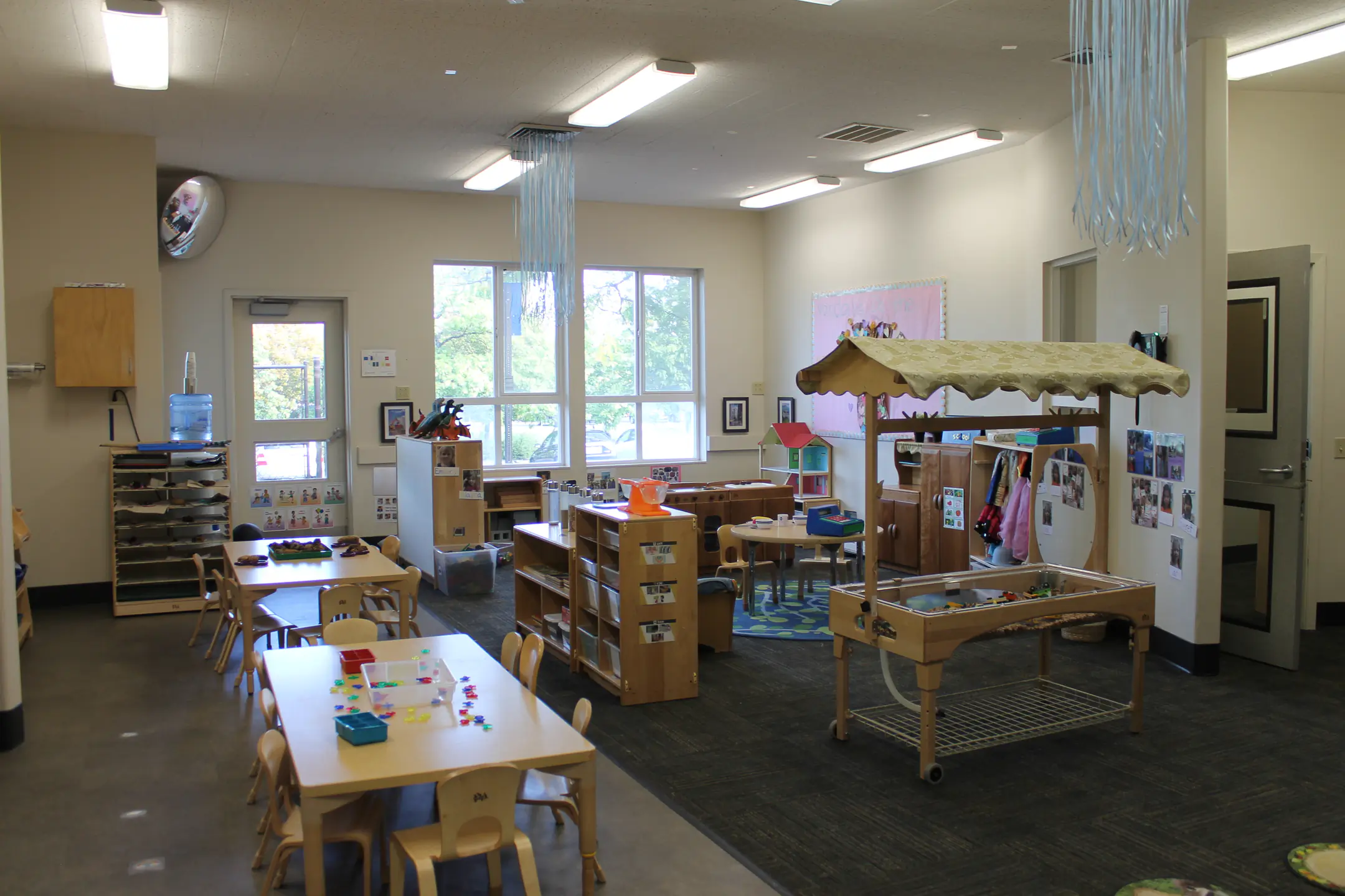 A classroom with group tables, sensory table, reading corner, and play area.