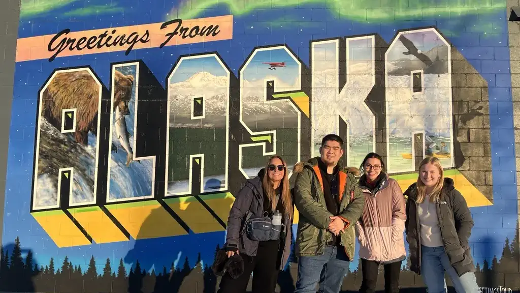 Four people pose against a painted wall mural that says “Greetings from Alaska.” 