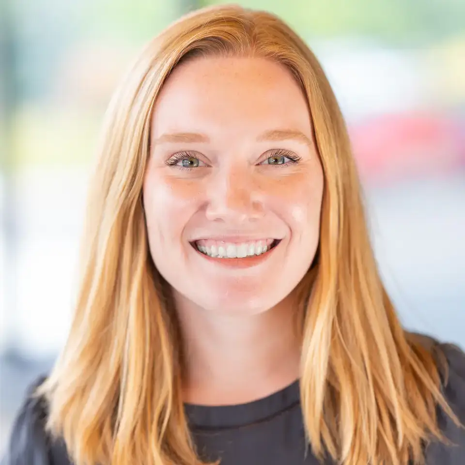 Smiling individual with shoulder-length blonde hair, wearing a black shirt, against a blurred outdoor background.