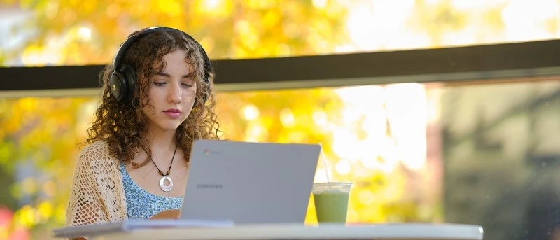 A female student works on a laptop at an outdoor table in the fall. 