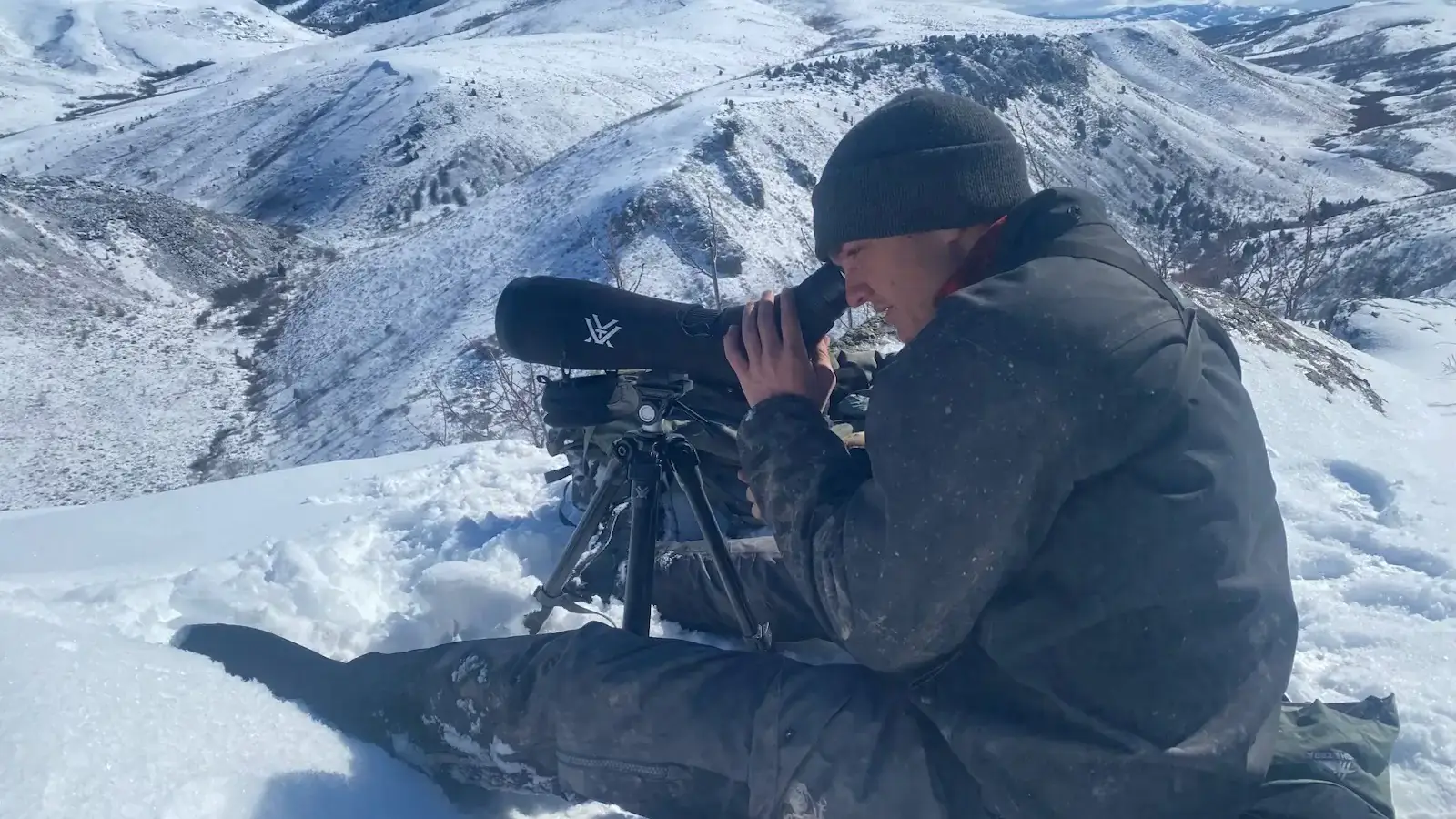 A man in an arctic setting looks through a telescope device to observe something in the distance.  