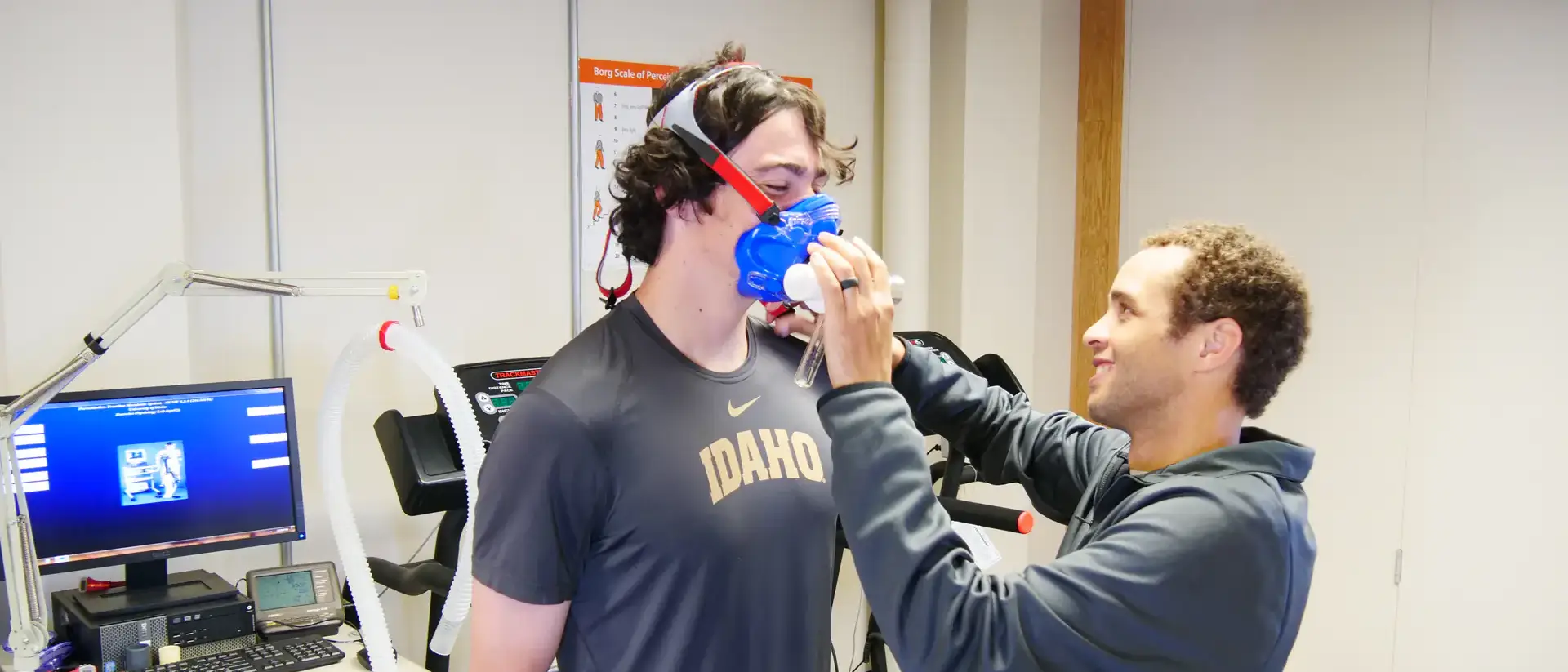 A person is being fitted with a blue respiratory testing mask by a technician in a lab with equipment and a treadmill.